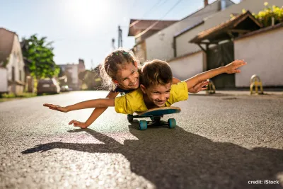 A happy small boy and a girl lying on a skateboard with arms outstreched as though flying. They are on the road, there are no cars and the sun is shining.