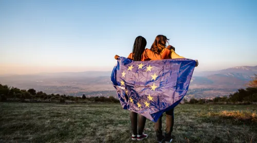Two women standng in remote landscape with EU flag wrapped around them
