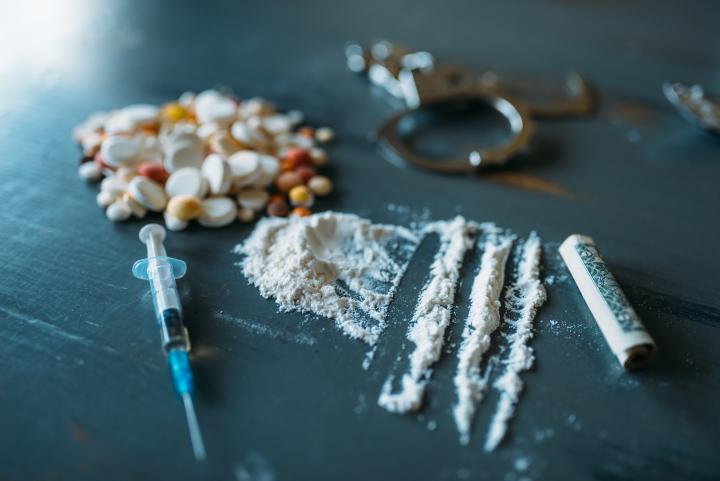 Close shot of different items laying in a surface: some drugs (cocaine and pills) and a syringe, also a pair of handcuffs in the background, more blurry. 
